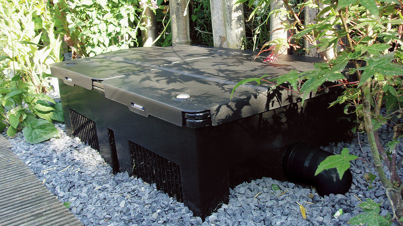 Das Bild zeigt den Oase BioSmart 36000 Durchlauffilter, ein hochwertiges Filtersystem für Gartenteiche. Der schwarze Filter ist rechteckig und steht auf einer Schicht aus grauem Kies, umgeben von üppigem Grün. Er ist für Teichbesitzer geeignet, die eine e