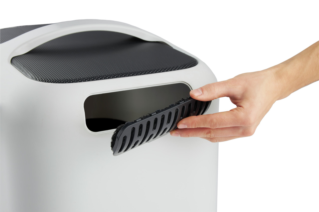 Das Bild zeigt eine weiße Katzentoilette der Marke Rotho mit einem modernen Design. Eine Hand entfernt gerade den schwarzen Aktivkohlefilter aus der Öffnung an der Vorderseite. Der Filter dient zur Geruchsneutralisierung. Die Oberfläche der Toilette ist g