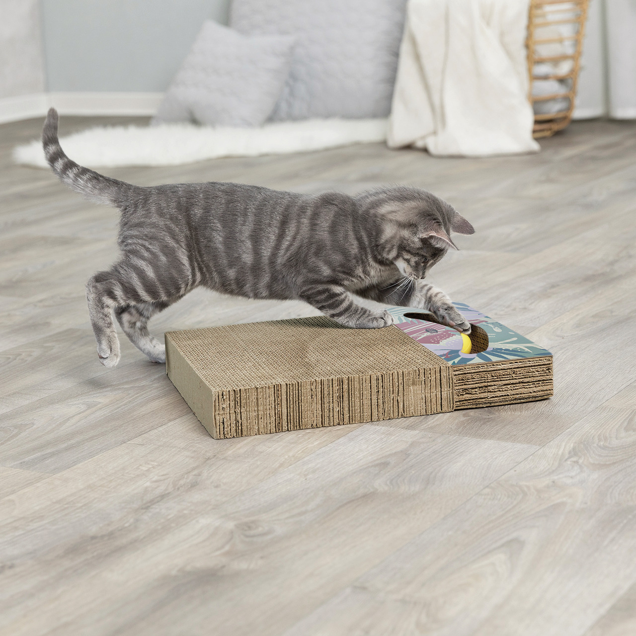 Das Bild zeigt ein TRIXIE Kratzbrett für Katzen, geeignet für junge und erwachsene Katzen. Es besteht aus Wellpappe und bietet eine integrierte Spielmöglichkeit mit buntem Design. Eine graue Katze spielt aktiv darauf, was die Beschäftigung und Krallenpfle