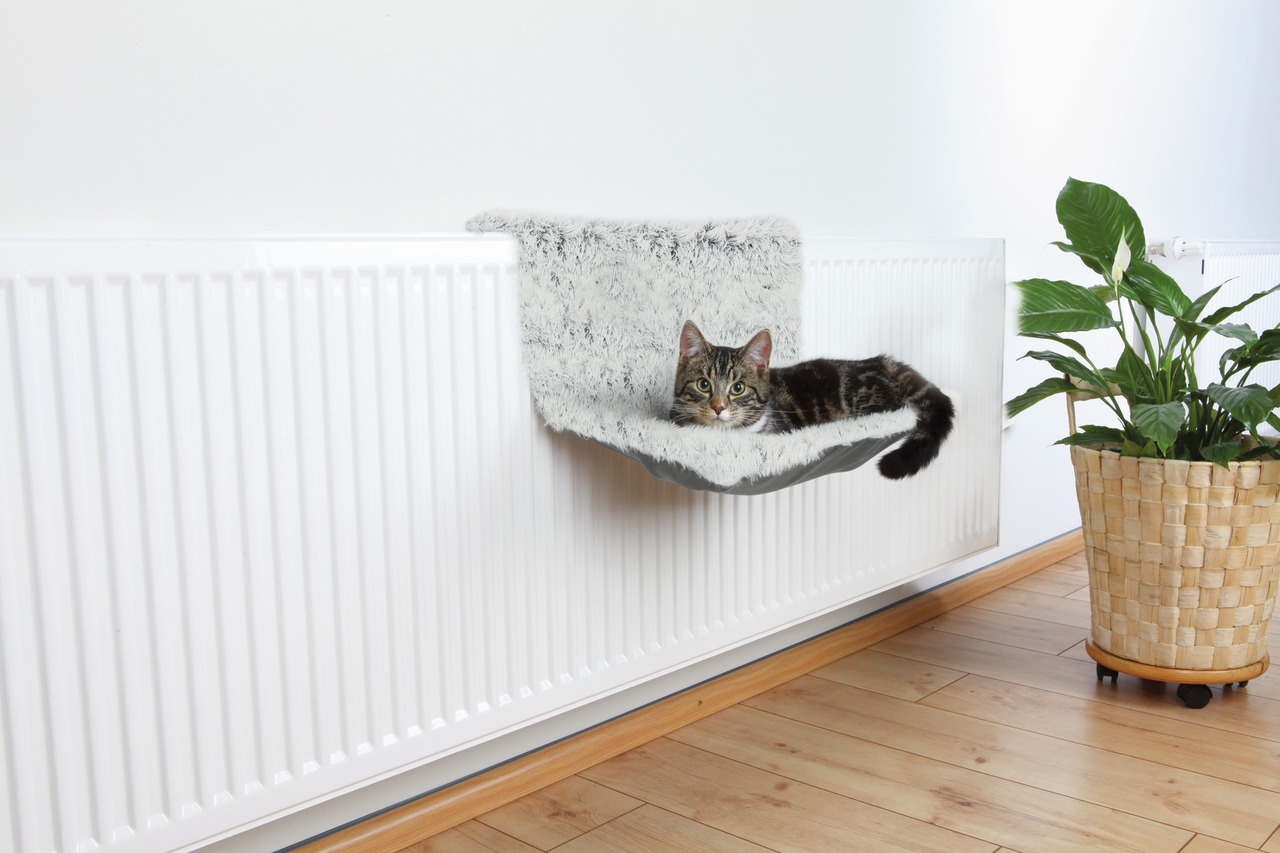 Das Bild zeigt eine Katze, die entspannt in einer TRIXIE Heizkörperliege liegt. Die Liege ist mit weichem, grauem Plüsch bezogen und speziell für Katzen entwickelt. Sie wird an einem weißen Heizkörper befestigt und bietet Wärme und Komfort. Im Hintergrund