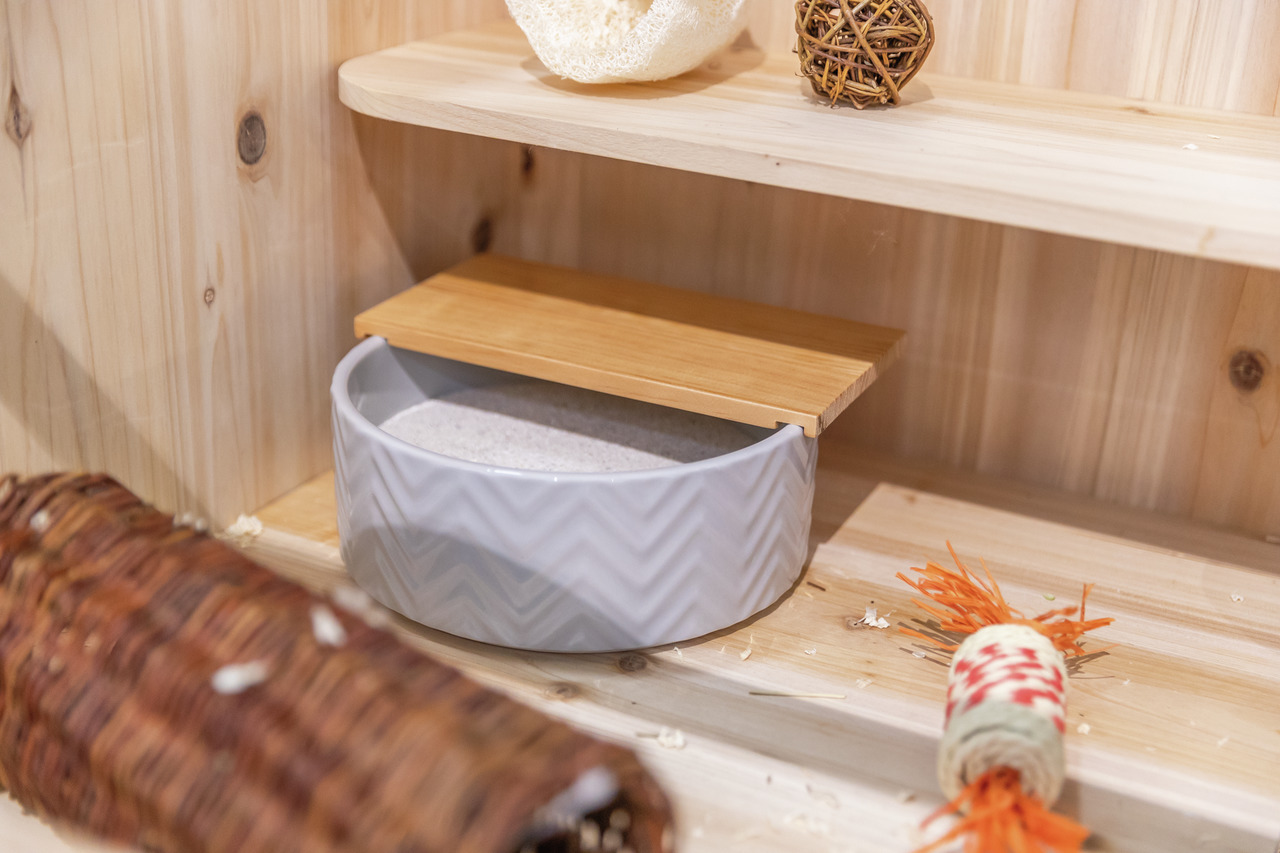 Das Bild zeigt eine TRIXIE Sandbad-Schale für Kleintiere, ideal für Hamster oder Gerbils. Die Schale besteht aus grauem Keramik mit Zickzack-Muster und einem abnehmbaren Holzdeckel. Sie ist mit feinem Sand gefüllt und steht in einem Holzgehege, umgeben vo