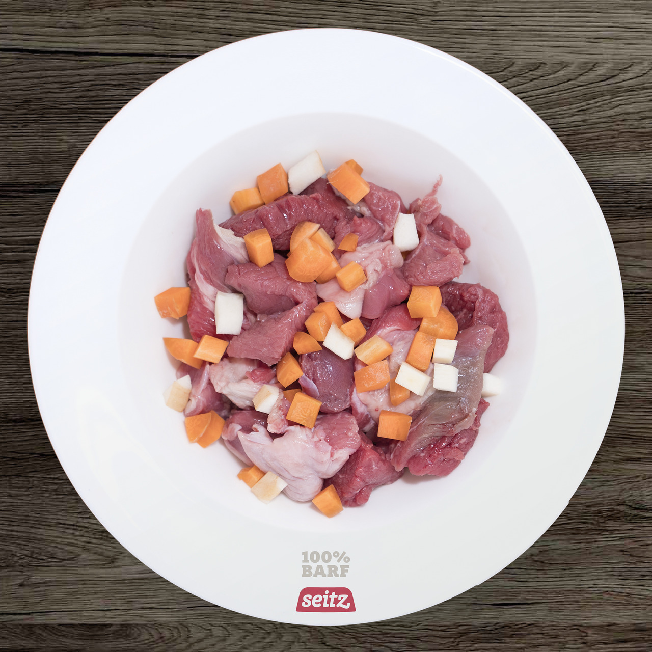 Das Bild zeigt eine weiße Schale mit frischem BARF-Hundefutter der Marke Seitz. Das Futter besteht aus rohem Fleisch, Karottenstücken und weißen Gemüsewürfeln. Es ist für Hunde geeignet und bietet eine natürliche, artgerechte Ernährung. Auf der Schale ste