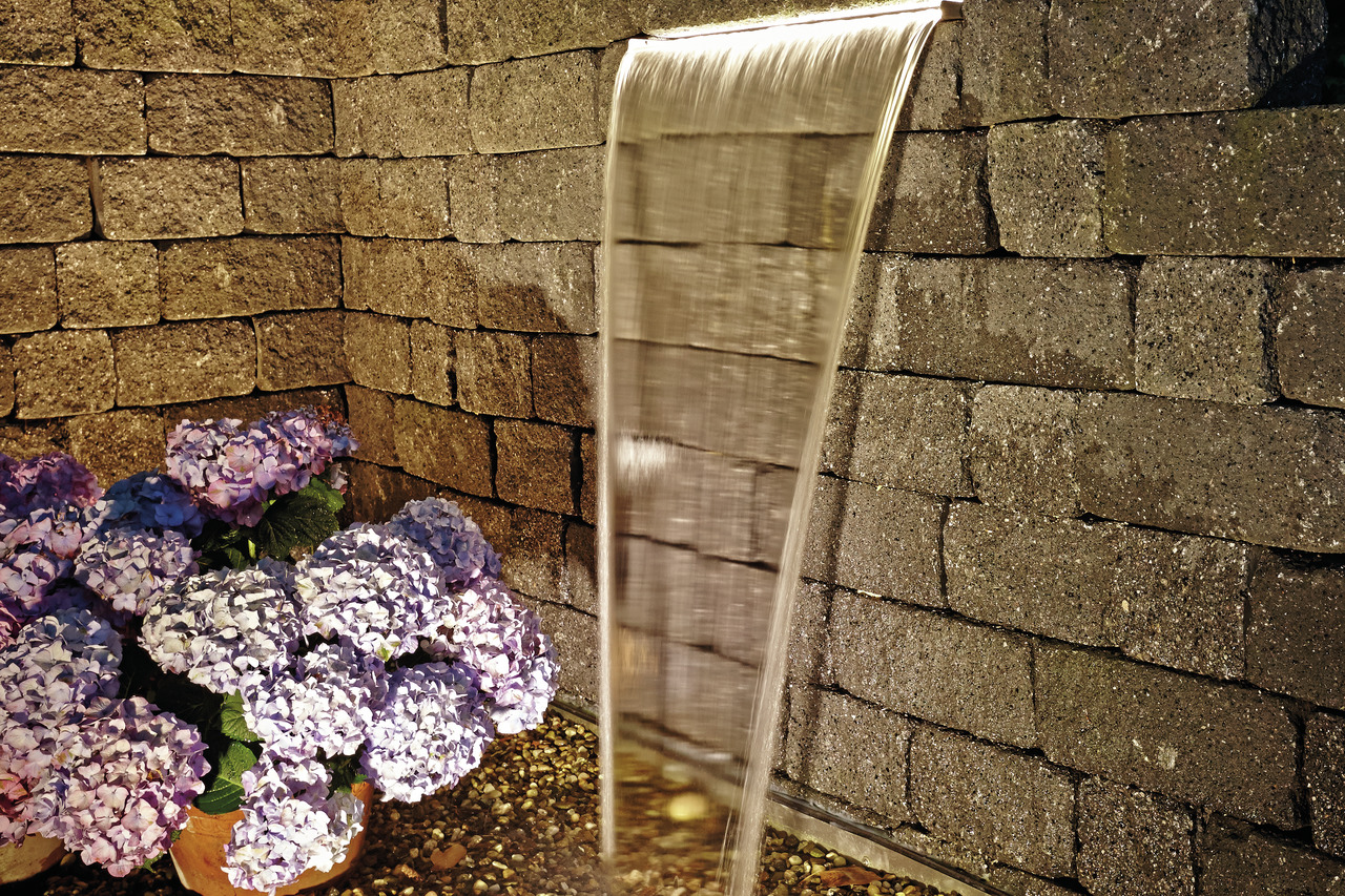 Oase Wasserfall, beleuchtete Kante, beruhigende Atmosphäre, Garten-Deko mit Hortensien in Violett/Blau, Natursteinmauer, Kiesboden.