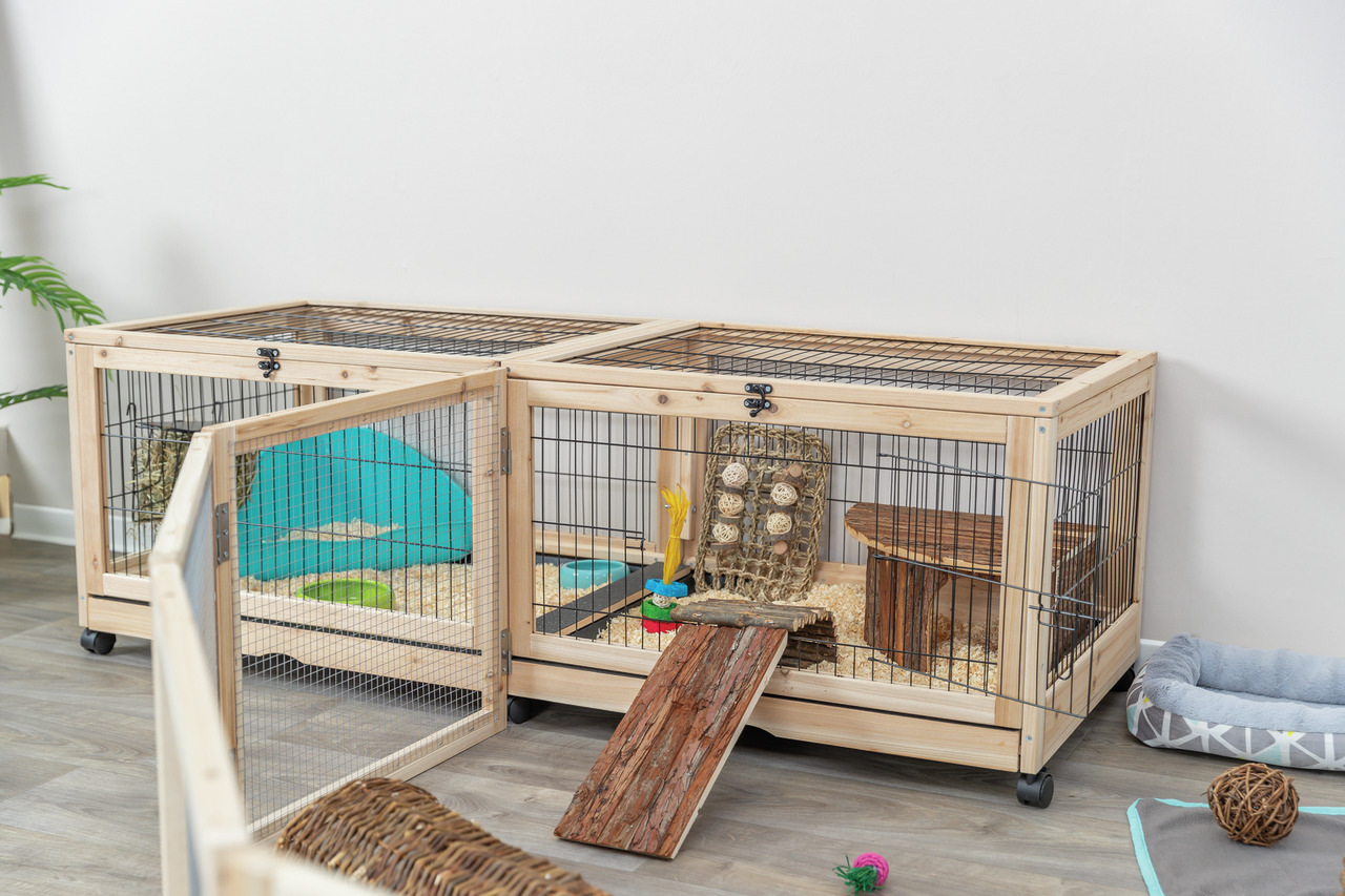Das Bild zeigt den TRIXIE Kleintierkäfig für Nager, geeignet für Kaninchen oder Meerschweinchen. Der Käfig besteht aus Holz und Metallgittern, verfügt über eine aufklappbare Tür und ist mit Rollen ausgestattet. Innen sind Spielzeuge, eine Rampe und Einstr