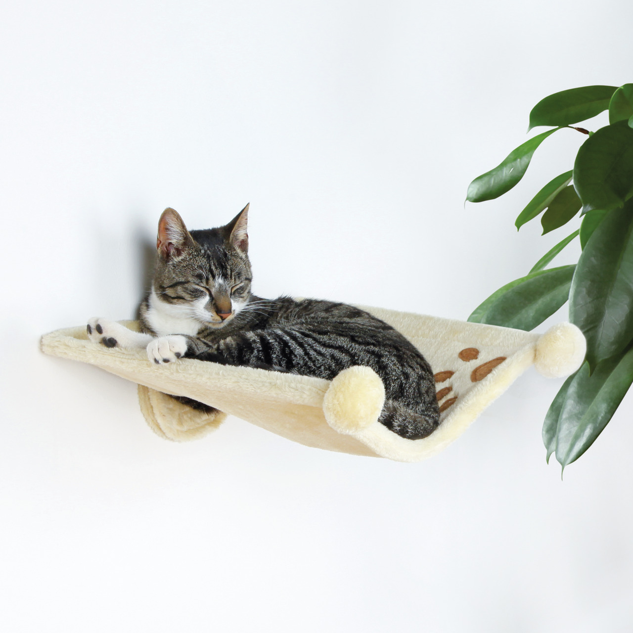 Das Bild zeigt eine grau-weiß getigerte Katze, die entspannt in einer cremefarbenen Wandliege von TRIXIE ruht. Die Liege ist mit weichem Plüsch bezogen und hat dekorative Pfotenabdrücke. Sie ist speziell für Katzen entwickelt und bietet eine gemütliche Rü