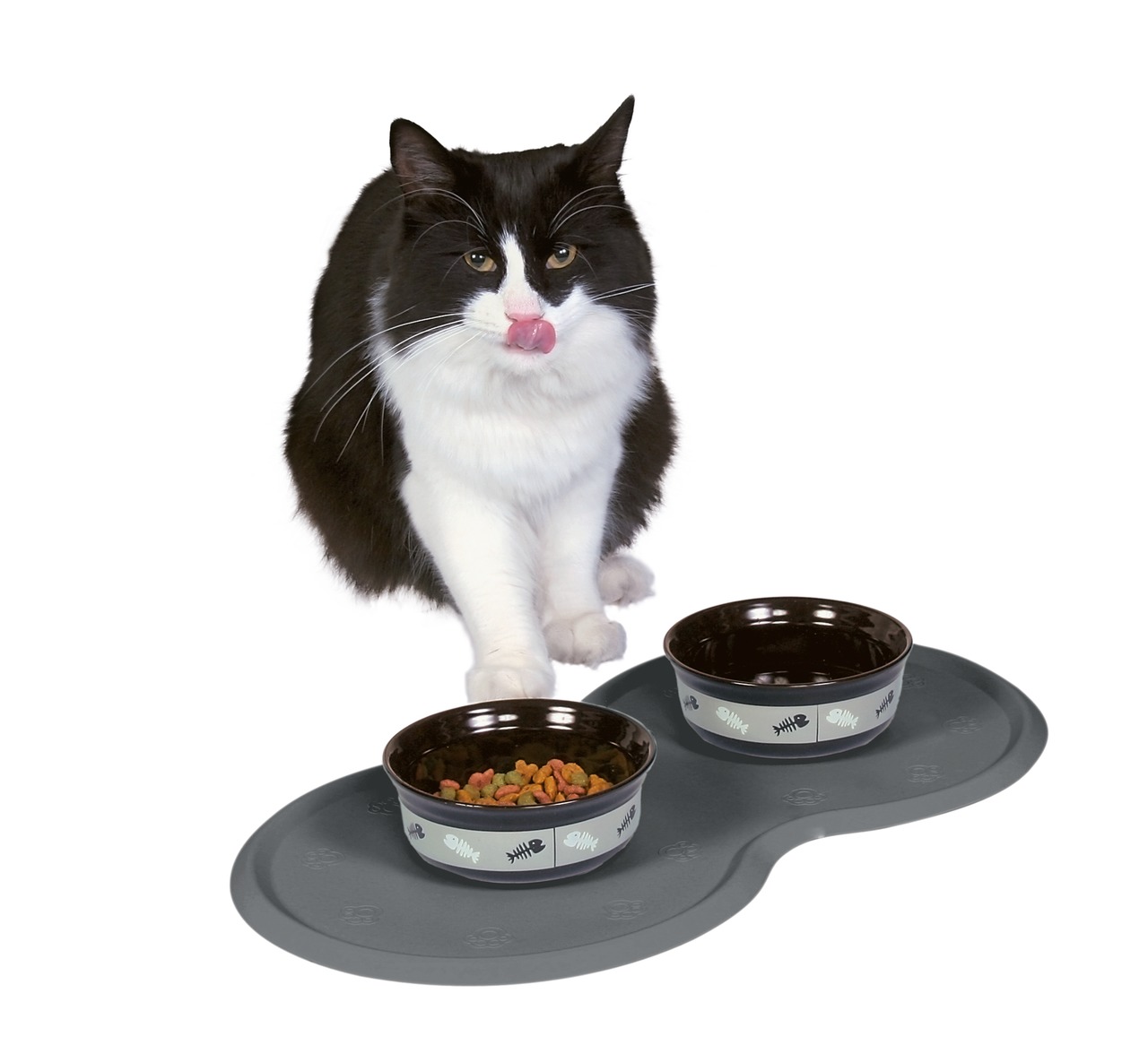Das Bild zeigt eine schwarz-weiße Katze, die vor zwei Futternäpfen auf einer grauen Unterlage sitzt. Die Näpfe gehören zum Produkt „TRIXIE Keramiknapf-Set mit Unterlage“ und sind für Katzen geeignet. Einer der Näpfe ist mit Trockenfutter gefüllt. Die Unte