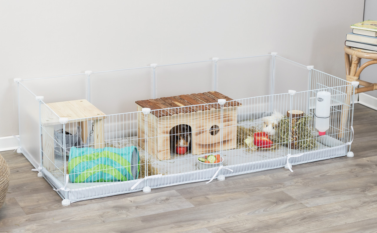 Das Bild zeigt den TRIXIE Kleintier-Freilauf aus weißem Metallgitter, geeignet für Kleintiere wie Meerschweinchen. Im Gehege befinden sich ein Holzhaus, ein Tunnel, eine Trinkflasche, Heu, Gemüse und ein Futternapf. Zielgruppe sind Kleintierhalter. Perfek