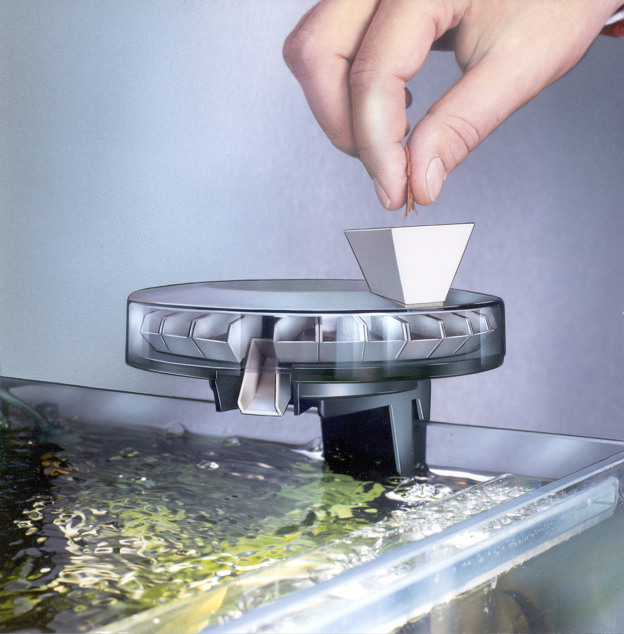Das Bild zeigt den Grässlin Rondomatic 400, einen automatischen Futterspender für Aquarien. Eine Hand füllt Futter in den Spender, der über einem Aquarium positioniert ist. Der Futterspender ist ideal für Aquarienfische und ermöglicht präzise Fütterung. K