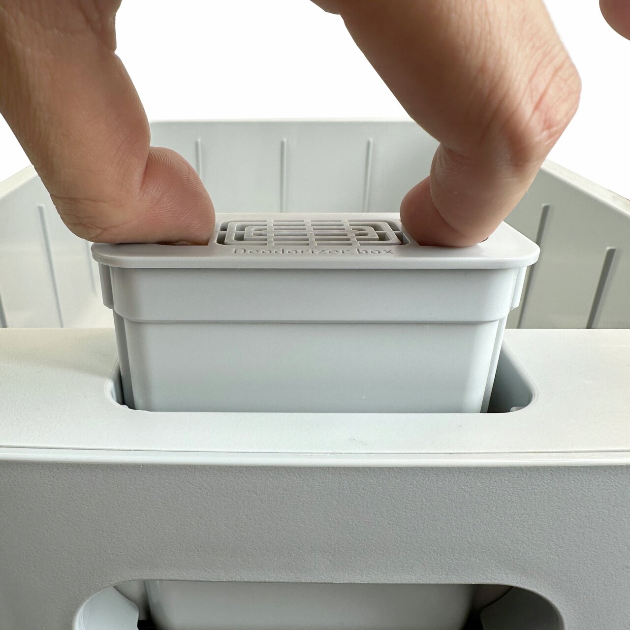 Ein weißer Filterbehälter wird in eine vorgesehene Öffnung eingesetzt. Der Text "Deodorizer box" ist auf dem Behälter sichtbar.