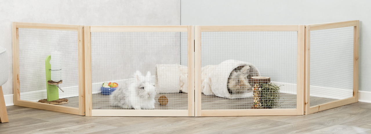 Das Bild zeigt ein Freigehege der Marke TRIXIE, geeignet für Kleintiere wie Kaninchen oder Meerschweinchen. Es besteht aus einem stabilen Holzrahmen mit Drahtgitter und bietet eine sichere Umgebung. Im Gehege befinden sich ein weißes Kaninchen, ein Tunnel