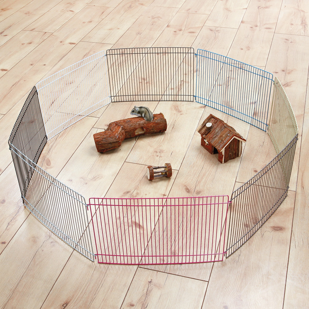 Das Bild zeigt den TRIXIE Freilaufgehege für Kleintiere, geeignet für Hamster und andere kleine Nager. Es besteht aus mehreren Metallgittern in verschiedenen Farben, die zu einem Kreis verbunden sind. Im Inneren befinden sich Zubehörteile wie ein Holzhaus