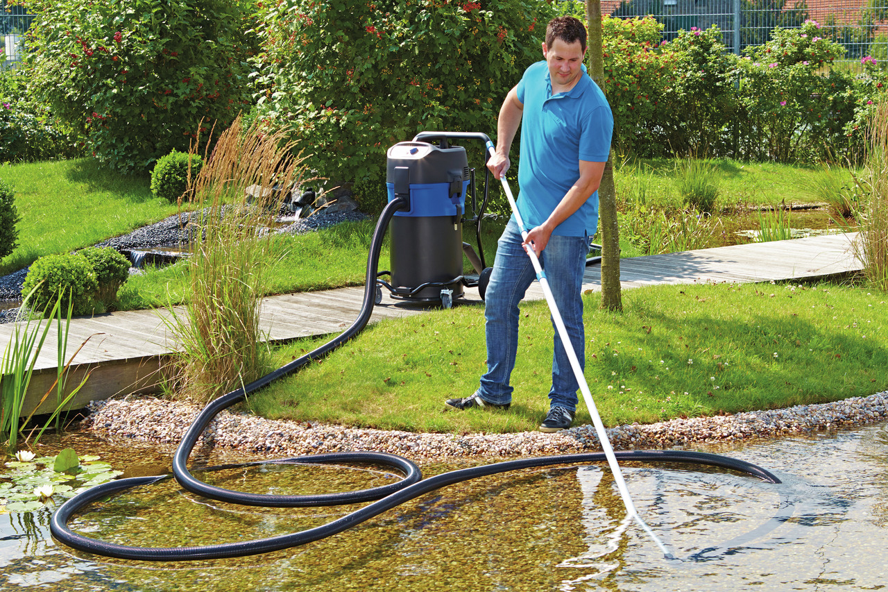 Ein Mann reinigt einen Gartenteich mit einem Oase Teichsauger. Der Oase PondoVac 4, ein leistungsstarker Teich- und Poolsauger, ist im Einsatz. Im Hintergrund sind ein gepflegter Garten mit Rasen, Sträuchern und ein Holzsteg zu sehen. Der Sauger ist mit e