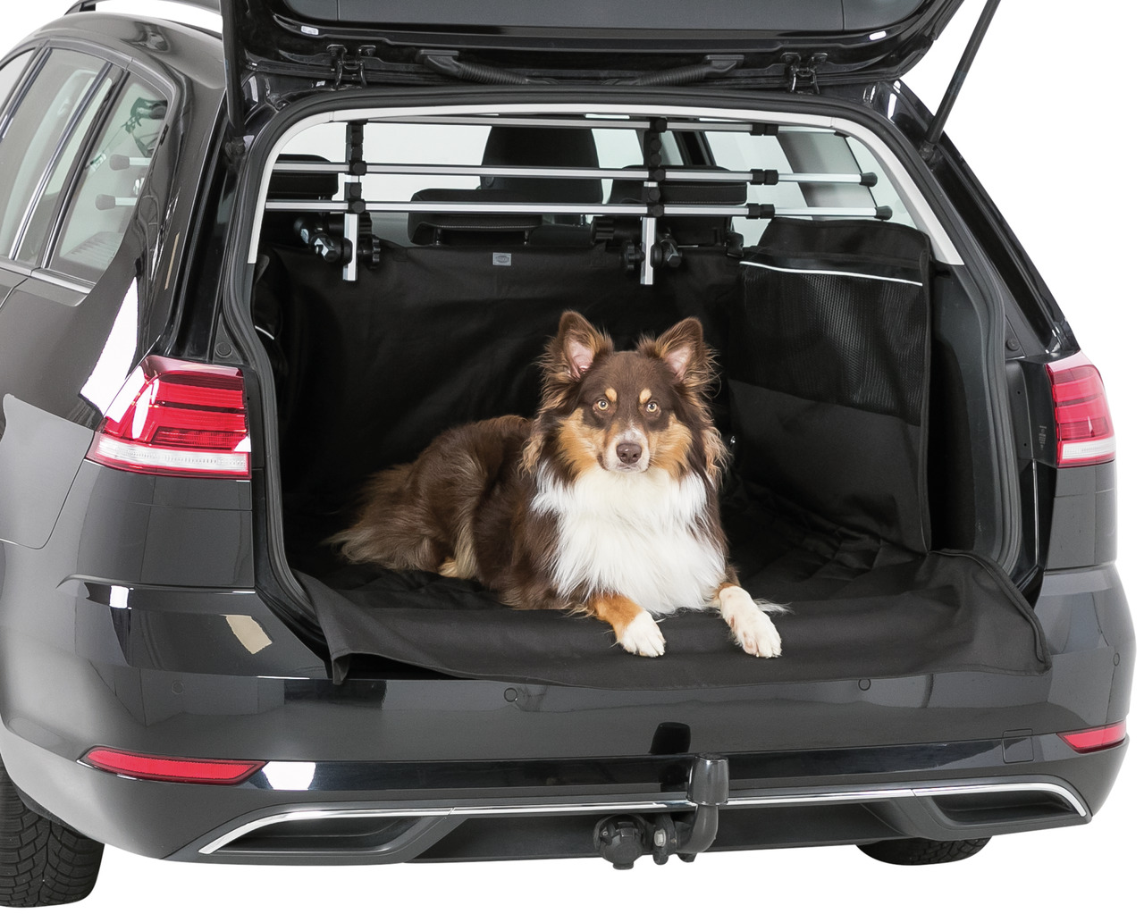 Das Bild zeigt einen braun-weißen Hund, der entspannt im Kofferraum eines Autos liegt. Der Kofferraum ist mit einer schwarzen Schutzdecke der Marke TRIXIE ausgekleidet, die speziell für Hunde entwickelt wurde. Die Schutzdecke schützt den Innenraum vor Sch