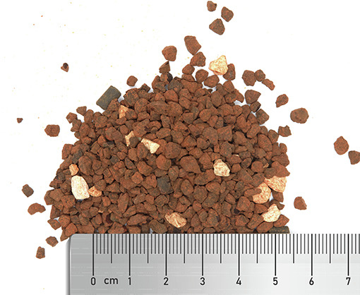 Ein Haufen braun-rötlicher Granulatstücke, teilweise mit helleren Einschlüssen, liegt auf einer weißen Oberfläche. Im Vordergrund ist ein Lineal zu sehen, das die Größe der Körner anzeigt. Das Produkt ist von der Marke FAUNA MARIN und dient als spezielles
