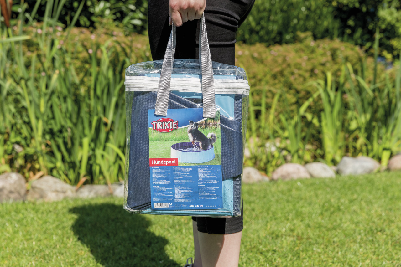 Eine Person hält eine transparente Tragetasche mit einem TRIXIE Hundepool. Die Verpackung zeigt ein Bild eines Hundes, der im blauen Pool sitzt. Der Pool ist für Hunde geeignet, bietet Abkühlung an heißen Tagen und ist leicht aufzubauen. Zielgruppe: Hunde