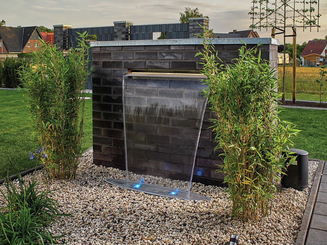 Das Bild zeigt einen modernen Gartenbrunnen der Marke Oase, der in eine stilvolle Steinmauer integriert ist. Der Wasserfall fließt über eine Edelstahlkante und wird von LED-Lichtern beleuchtet, die für eine stimmungsvolle Atmosphäre sorgen. Umgeben von Ba