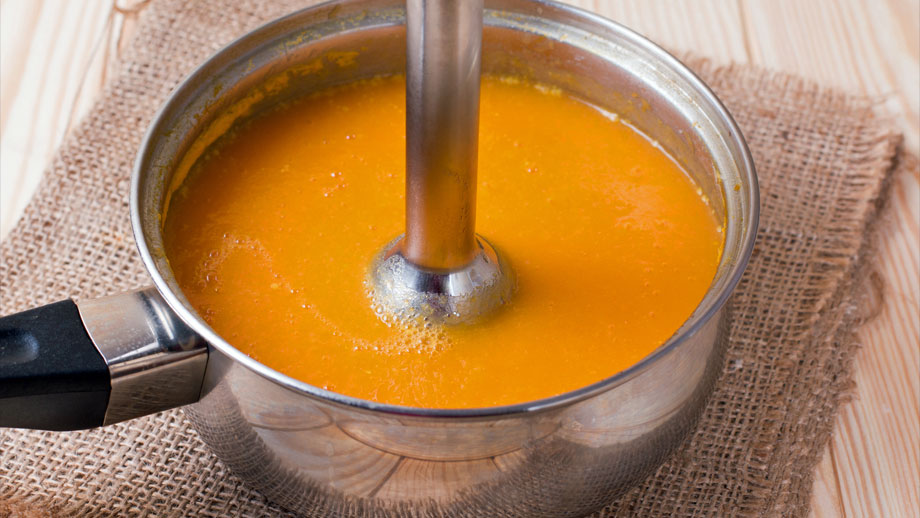 Karottensuppe in silbernem Topf wird mit Stabmixer püriert.