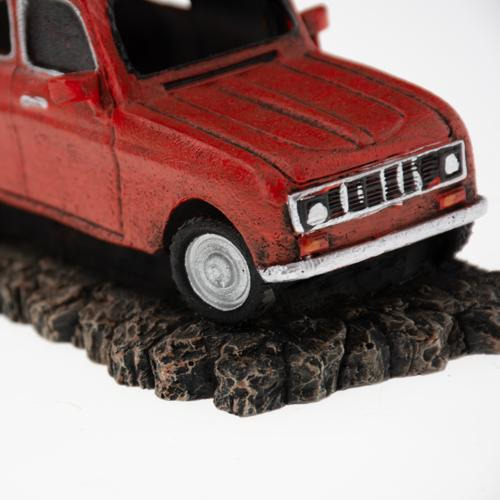 Ein rotes Modellauto, das auf einer felsigen, dunklen Oberfläche steht. Das Fahrzeug ist detailliert gestaltet, mit sichtbaren Scheinwerfern, Reifen und einer strukturierten Karosserie. Es handelt sich um ein dekoratives Element der Marke AQUA DELLA, geei