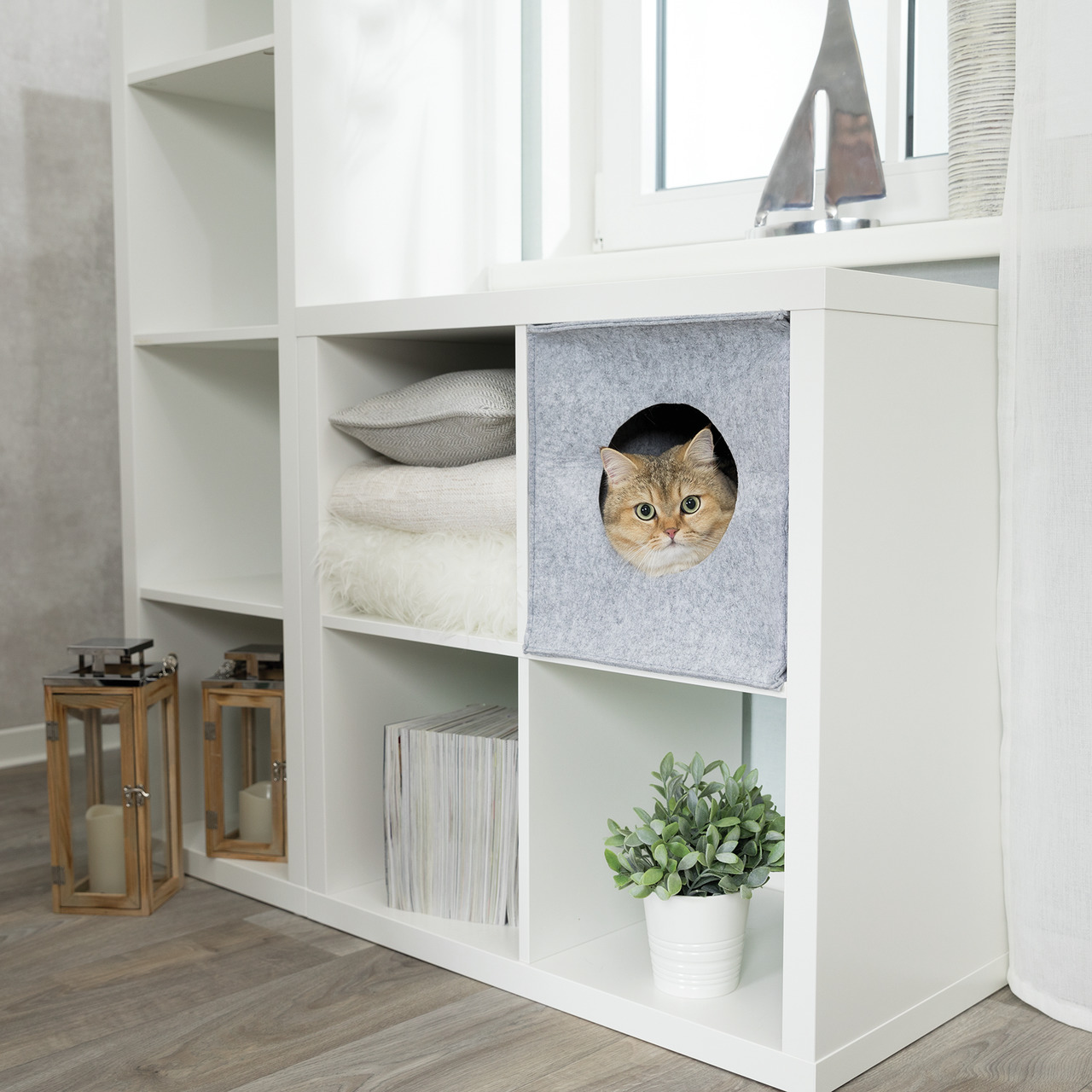 Auf dem Bild ist ein modernes Regal mit mehreren Fächern zu sehen. In einem der Fächer befindet sich die TRIXIE Kuschelhöhle für Katzen, geeignet für alle Katzenrassen. Die Höhle ist grau und aus Filz gefertigt, ideal für Rückzug und Entspannung. Daneben 