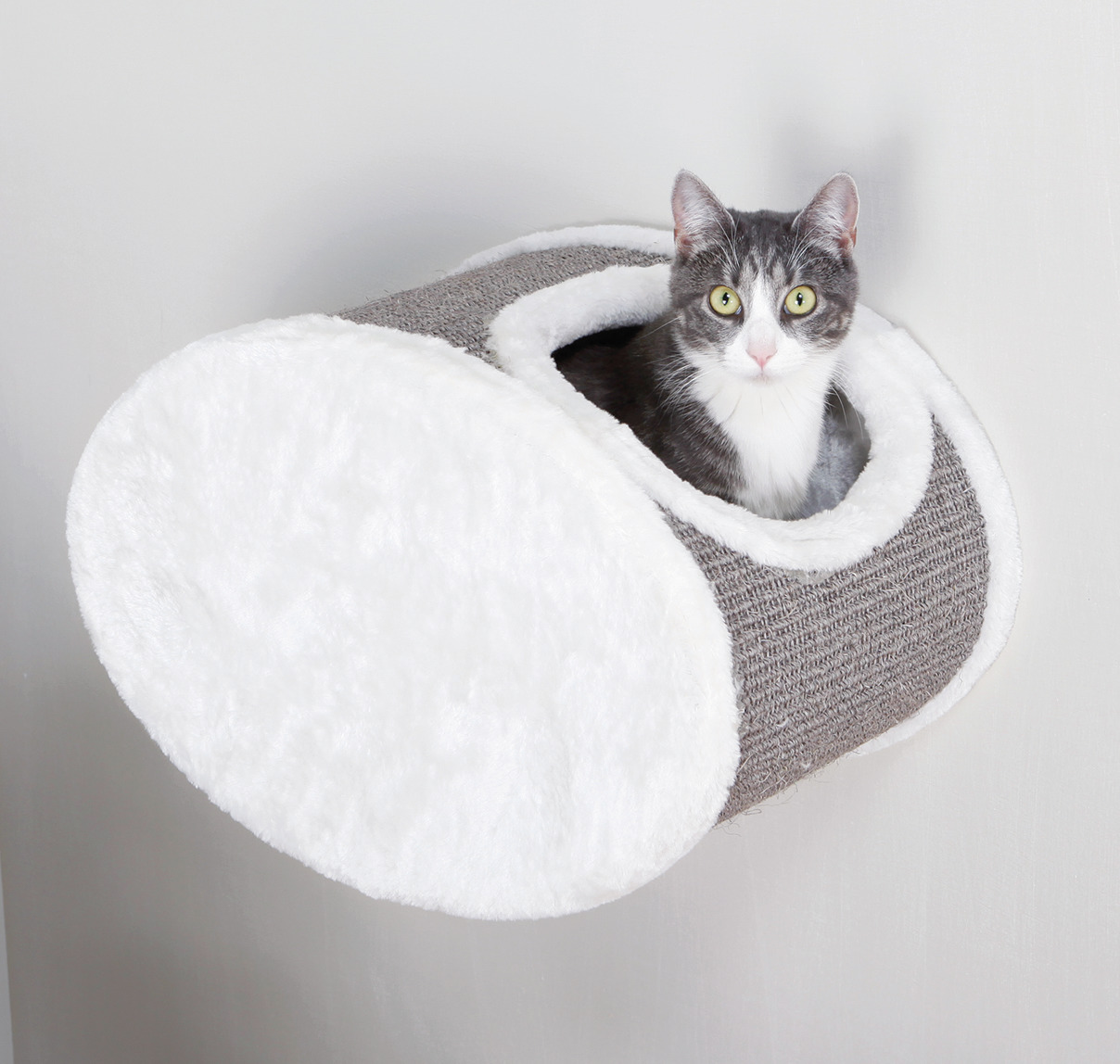 Das Bild zeigt eine grau-weiße Katze, die in einer TRIXIE-Wandliege für Katzen sitzt. Die Liege ist zylinderförmig, mit einer weichen weißen Plüschoberfläche und einer grauen Kratzfläche aus Sisal. Sie ist speziell für Katzen entwickelt und bietet eine ge