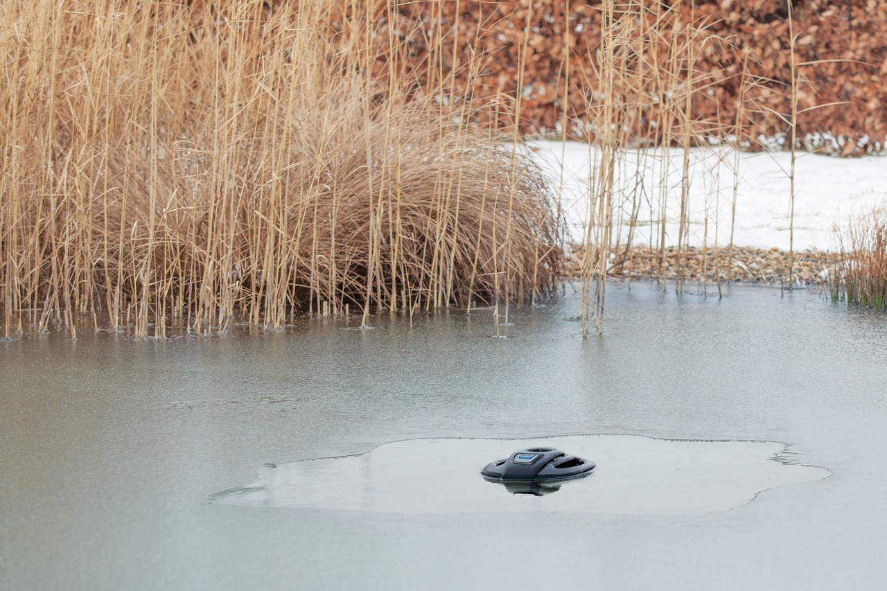 Das Bild zeigt den Oase IceFree 4 Seasons, ein hochwertiges Eisfreihalter-Gerät für Gartenteiche. Es schwimmt auf einer teilweise gefrorenen Wasseroberfläche und sorgt für eine eisfreie Zone, um den Gasaustausch im Winter zu gewährleisten. Im Hintergrund 