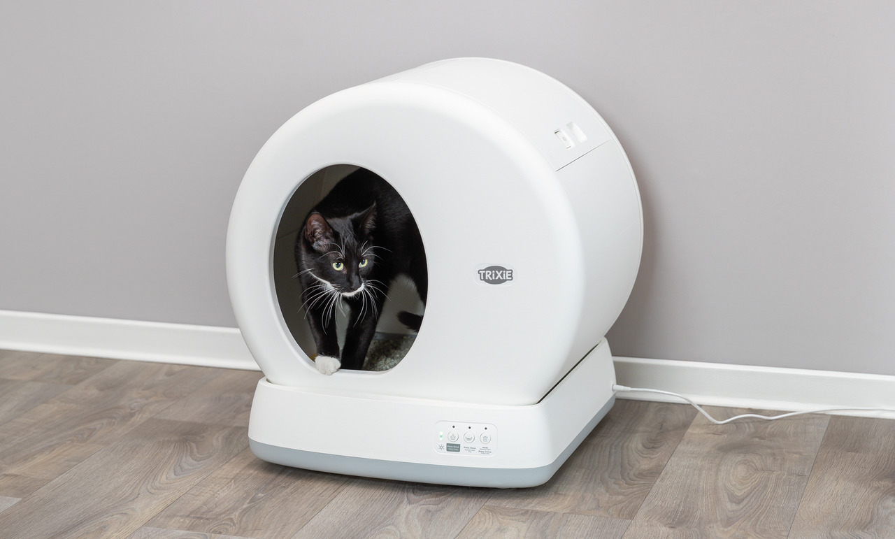 Das Bild zeigt eine automatische Katzentoilette der Marke TRIXIE, Modell "Smart Litter Box", geeignet für Katzen. Die weiße, runde Toilette steht auf einem Holzboden und ist mit einem schwarzen Kabel angeschlossen. Eine schwarz-weiße Katze schaut aus der 