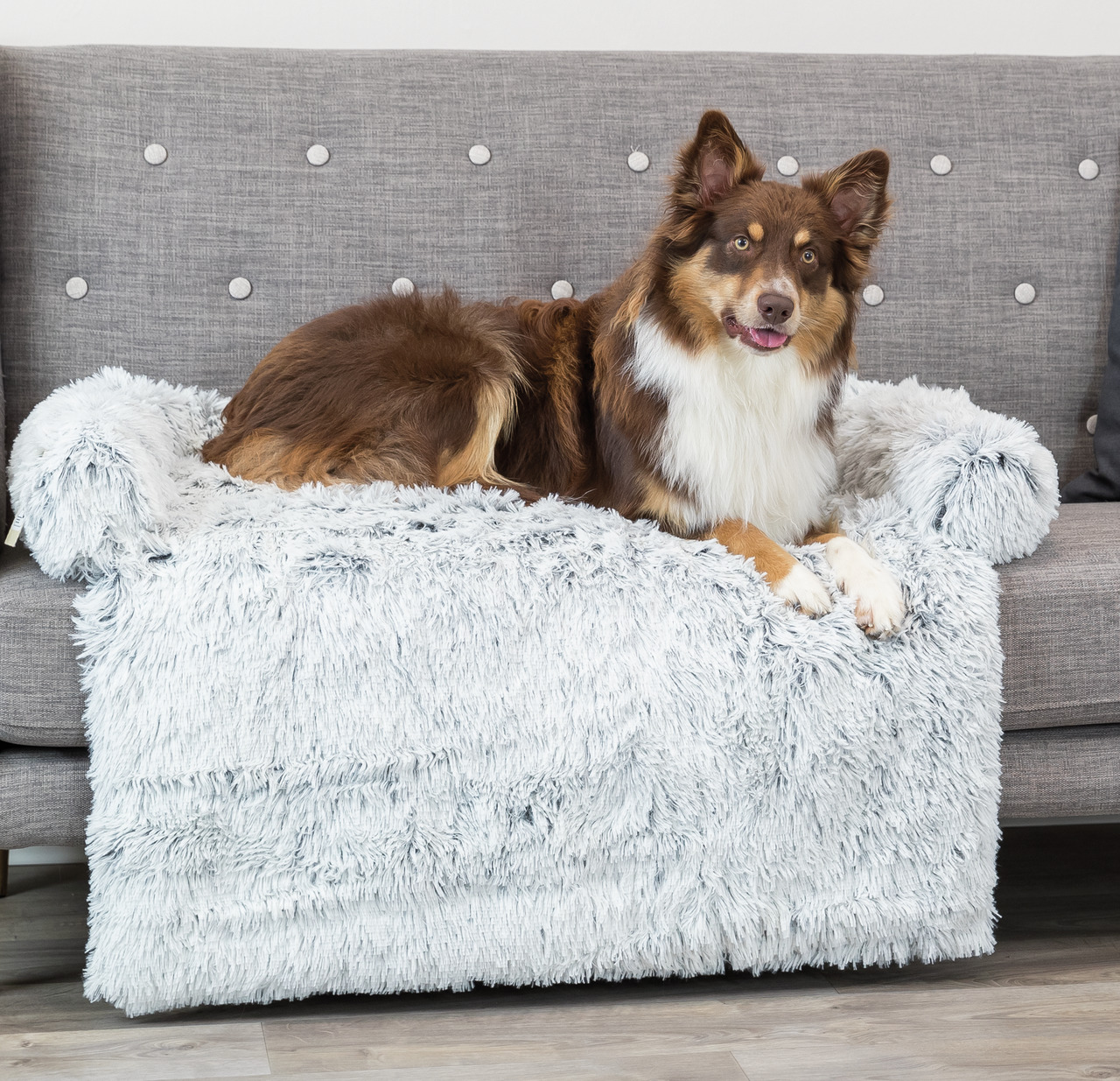 Ein braun-weißer Hund liegt entspannt auf einer grauen Couch, die mit einer flauschigen, weißen TRIXIE Sofadecke bedeckt ist. Die Decke ist speziell für Hunde konzipiert und bietet Komfort sowie Schutz für Möbel. Zielgruppe sind Hundebesitzer, die Wert au