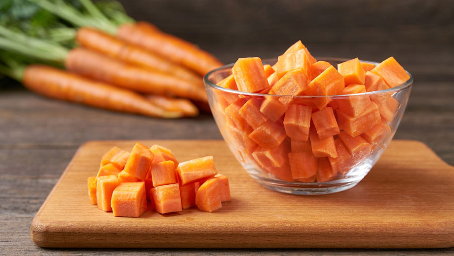 Würfel aus Karotten in Glasschüssel und auf Holzbrett, hinten Möhren mit Grün