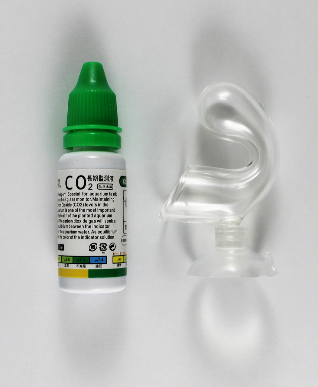 Das Bild zeigt das Produkt „Amazing Aqua CO2-Indikatorlösung“ für Aquarien. Links ist eine kleine weiße Flasche mit grünem Deckel und detaillierter Beschriftung zu sehen, die die CO2-Lösung enthält. Rechts daneben befindet sich ein transparenter Glasindik