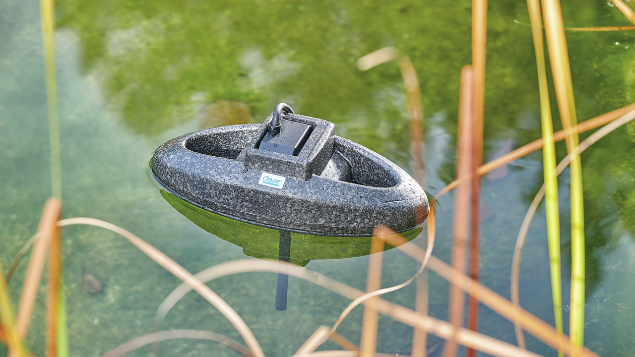 Schwimmender Teichbelüfter Oase AquaOxy 500, ideal für die Sauerstoffversorgung von Gartenteichen. Zielgruppe: Teichbesitzer. Das Bild zeigt das kompakte, graue Gerät auf der Wasseroberfläche eines Teiches, umgeben von Pflanzen. Fördert klares Wasser und 