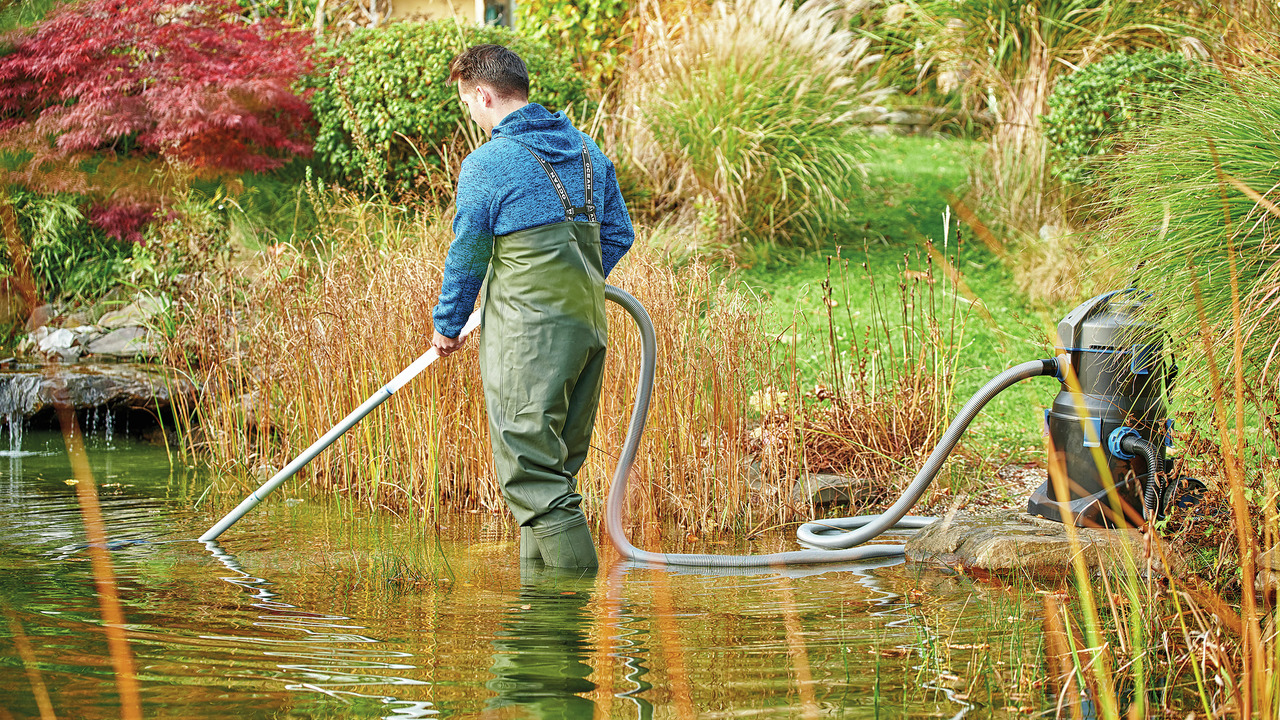 Ein Mann reinigt einen Gartenteich mit Oase Teichsauger: langes Saugrohr, flexibler Schlauch, gepflegte Gartenlandschaft im Hintergrund.