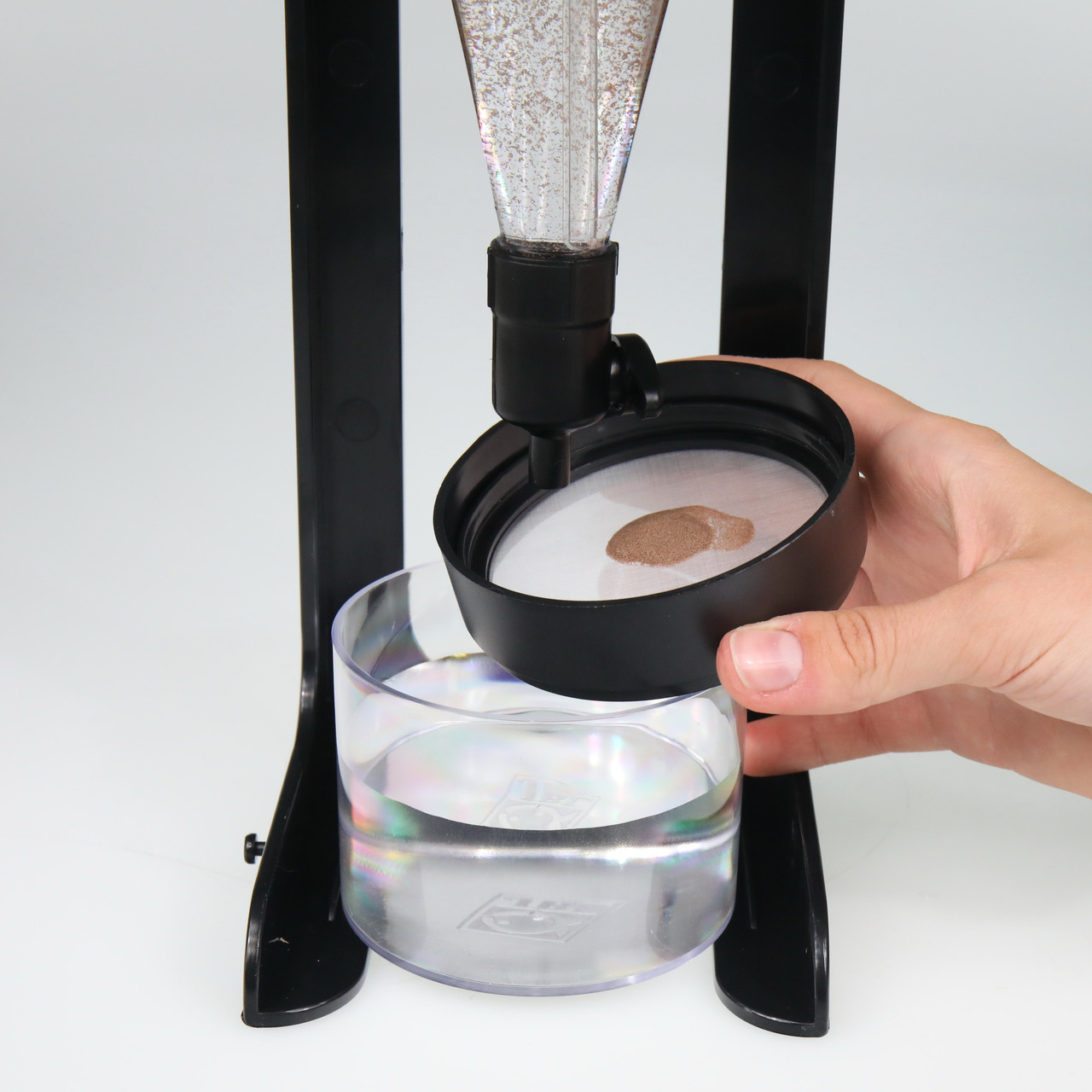 Das Bild zeigt das JBL ProScan Reagenzglas-Halterungssystem, ein Produkt für Aquaristik-Enthusiasten. Es wird verwendet, um Wasserproben präzise zu analysieren. Eine Hand hält den Deckel mit einem feinen Filter, während das Reagenzglas darunter positionie