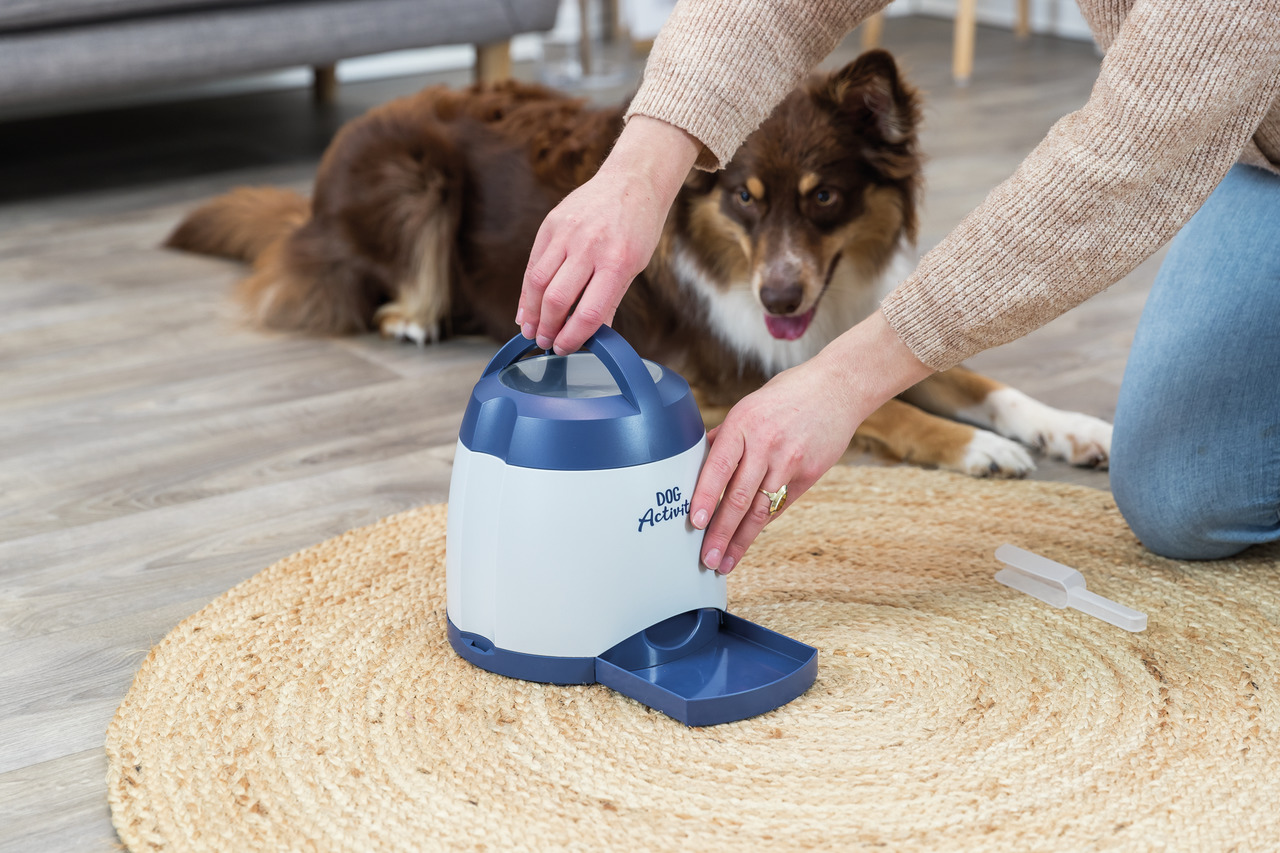 Das Bild zeigt den TRIXIE Dog Activity Snackball, ein interaktives Spielzeug für Hunde. Eine Person demonstriert die Nutzung, während ein Hund aufmerksam zuschaut. Das Produkt ist für Hunde aller Größen geeignet und dient der mentalen Stimulation. Es hat 