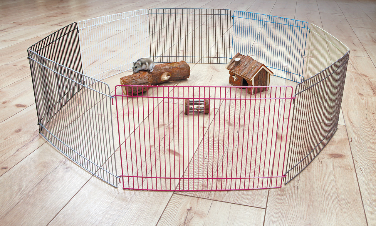Das Bild zeigt den TRIXIE Freilaufgehege für Kleintiere, geeignet für Hamster und andere kleine Nager. Das Gehege besteht aus mehreren Metallgittern in verschiedenen Farben, die zu einem Kreis verbunden sind. Im Inneren befinden sich ein Holzhaus, ein Hol