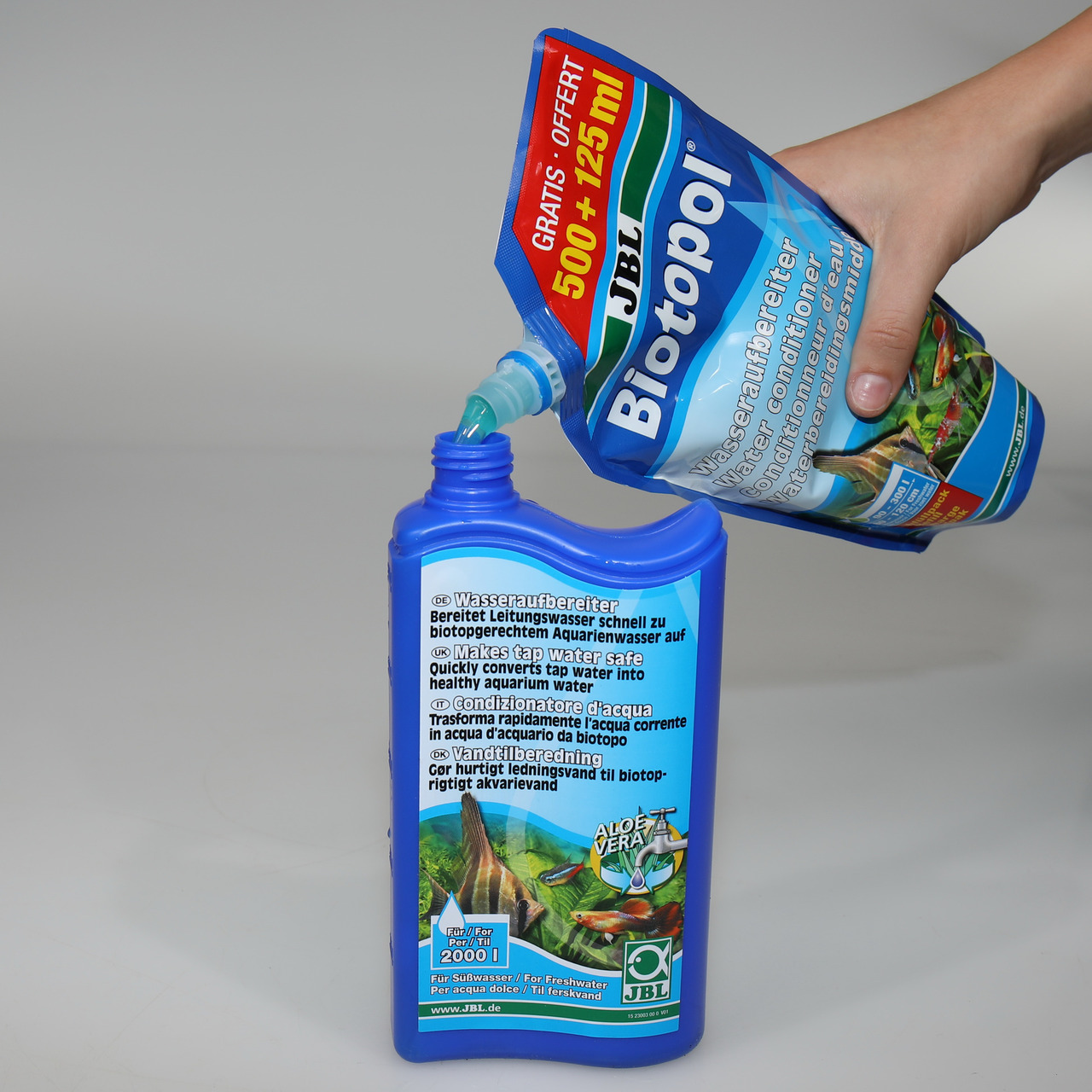 Das Bild zeigt eine Hand, die JBL Biotopol, einen Wasseraufbereiter für Aquarien, aus einer blauen Nachfüllpackung in eine blaue Flasche gießt. Das Produkt ist für Süß- und Teichwasser geeignet und enthält Aloe Vera. Die Verpackung weist auf eine Menge vo