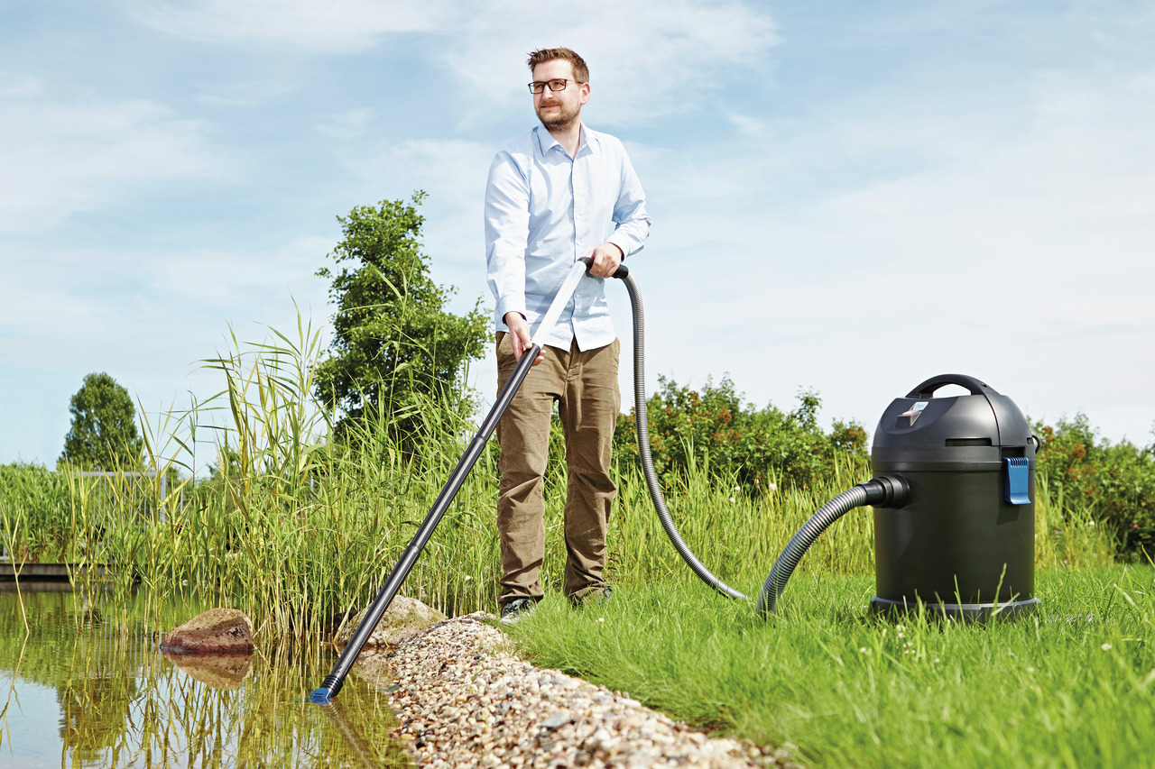 Ein Mann verwendet den Oase PondoVac 4, einen leistungsstarken Teich- und Poolsauger, um den Rand eines Gartenteichs zu reinigen. Das Gerät ist schwarz, mobil und mit einem flexiblen Saugschlauch ausgestattet. Im Hintergrund sind grüne Pflanzen und ein kl