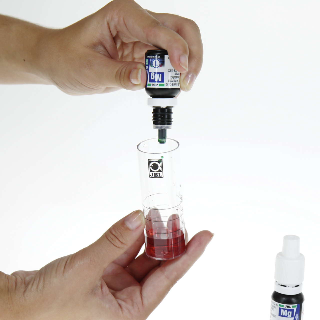 Das Bild zeigt eine Hand, die eine Flasche JBL Magnesium-Testlösung hält und Tropfen in einen transparenten Messbecher mit roter Flüssigkeit gibt. Der Messbecher trägt das JBL-Logo. Das Produkt ist für Aquarienbesitzer geeignet, um den Magnesiumgehalt im 