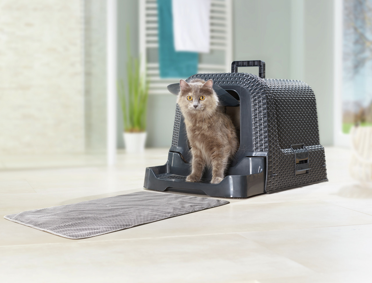 Das Bild zeigt eine graue Transportbox der Marke AniOne, geeignet für Katzen. Die Box hat ein modernes, geflochtenes Design und ist mit einer rutschfesten Matte ausgestattet, die vor der Box liegt. Eine graue Katze sitzt in der Box, die in einem hellen, w