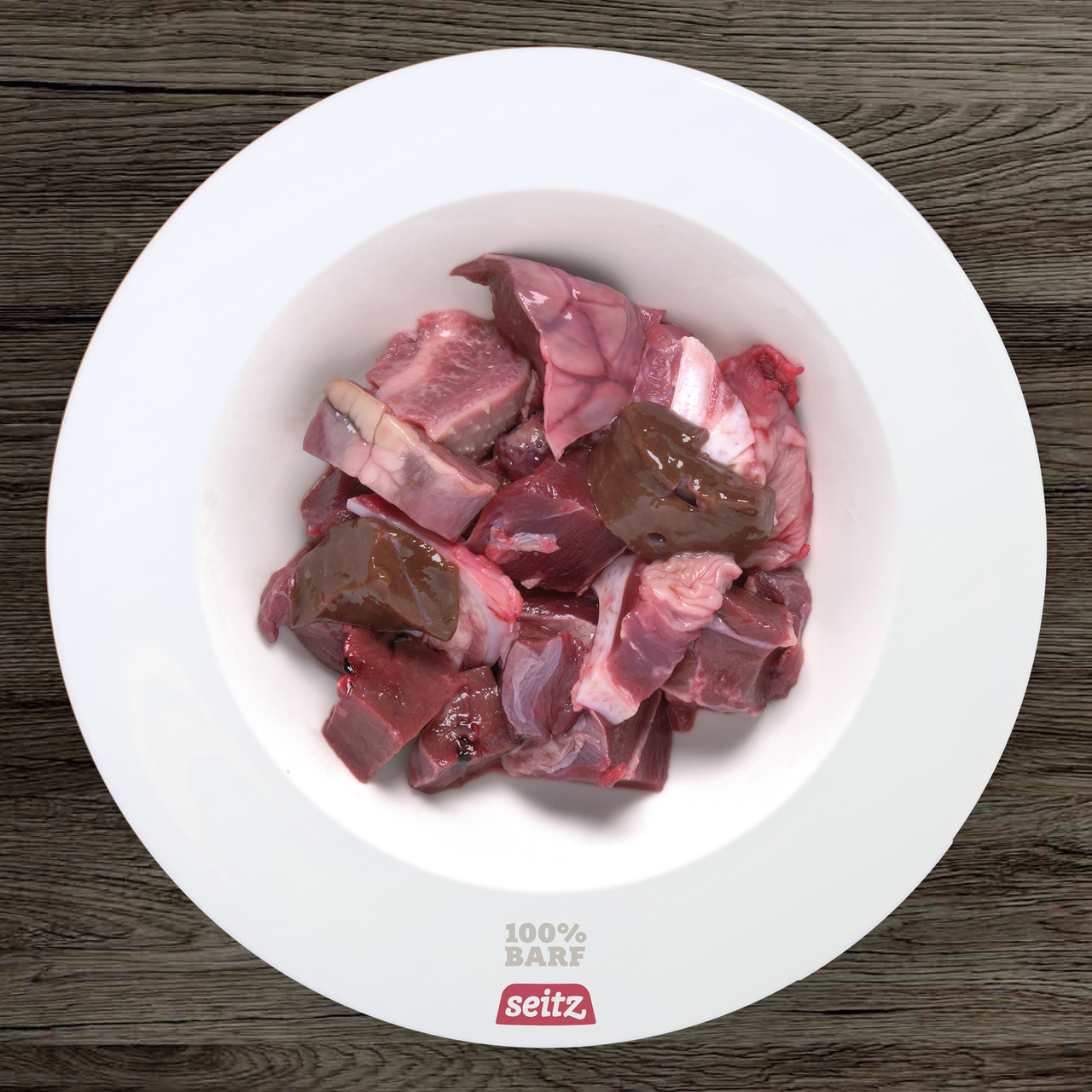 Das Bild zeigt eine weiße Futterschale mit rohem Fleisch, bestehend aus verschiedenen Fleischstücken und Innereien, die für die BARF-Ernährung von Hunden geeignet sind. Es handelt sich um das Produkt „Seitz 100% BARF“, speziell für Hunde. Die Marke Seitz 