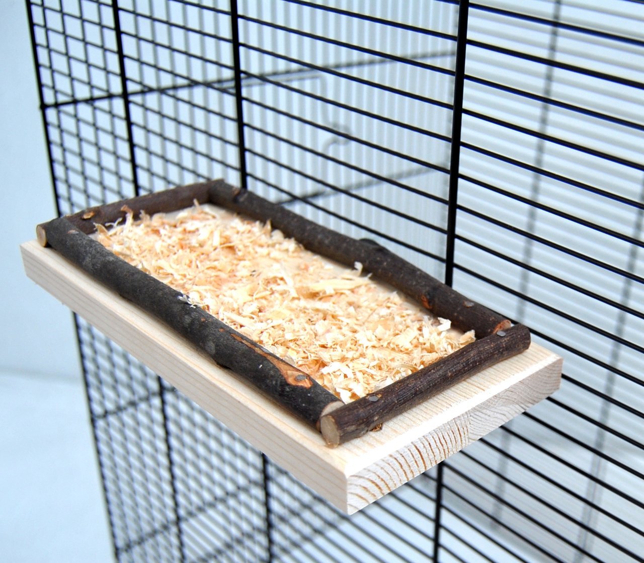 Das Bild zeigt eine natürliche Sitz- und Ruhefläche für Kleintiere, speziell für Vögel oder Nagetiere, der Marke Ollesch. Das Produkt besteht aus unbehandeltem Holz mit einem Rahmen aus Naturästen und einer Schicht aus Holzspänen. Es ist für Käfige geeign
