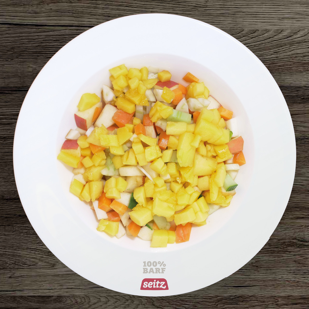 Das Bild zeigt eine weiße Schale mit einer Mischung aus frischen, gewürfelten Obst- und Gemüsestücken, darunter gelbe Mango, orange Karotten, grüne Gurken und weiße Apfelstücke. Auf der Schale steht „100% BARF“ und das Logo der Marke Seitz. Dieses Produkt